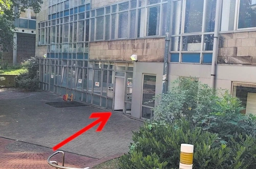 Die Treppen nach Rechts gehe bitte runter. Dort befindet sich auch eine Rampe. Wenn du die Treppen runtergegangen bist, findest du rechts ein Hochbeet mit Pflanzen. Wenn du dem folgst und dann der Wand, die sich im 90°-Winkel zum Beet befindest, findest du den Eingang zum Gebäude.